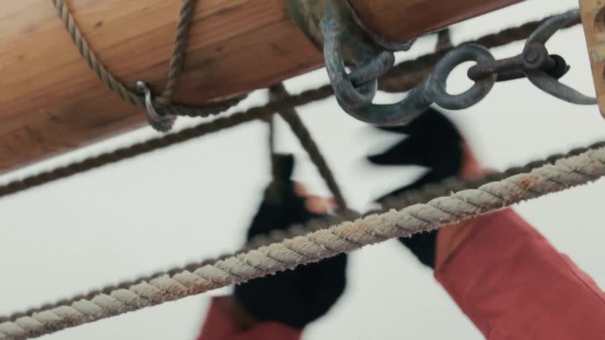 A sailor in a red jacket and gloves works with the ropes and rigging on the wooden mast of a ship