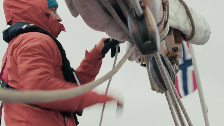 Dressed in a red rain jacket, a sailor skillfully secures the ropes on a classic wooden vessel with a Norwegian flag waving behind