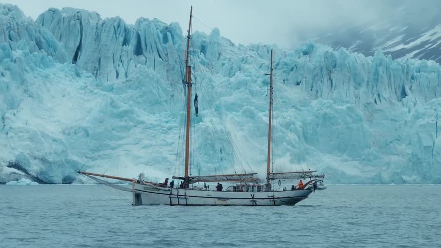 A majestic sailboat sits in the tranquil arctic waters as a massive glacier calves in the background