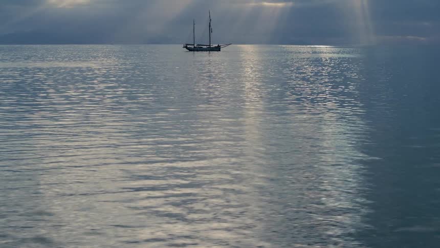 Sunbeams pierce through a dramatic cloudy sky, illuminating a lone schooner on a tranquil, glassy sea