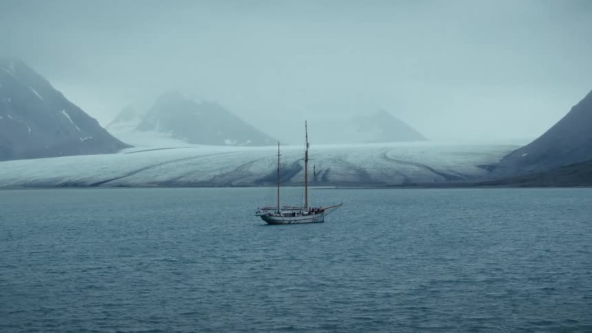A classic two-masted sailboat floats in the calm, misty waters of a remote arctic bay in front of a vast glacier