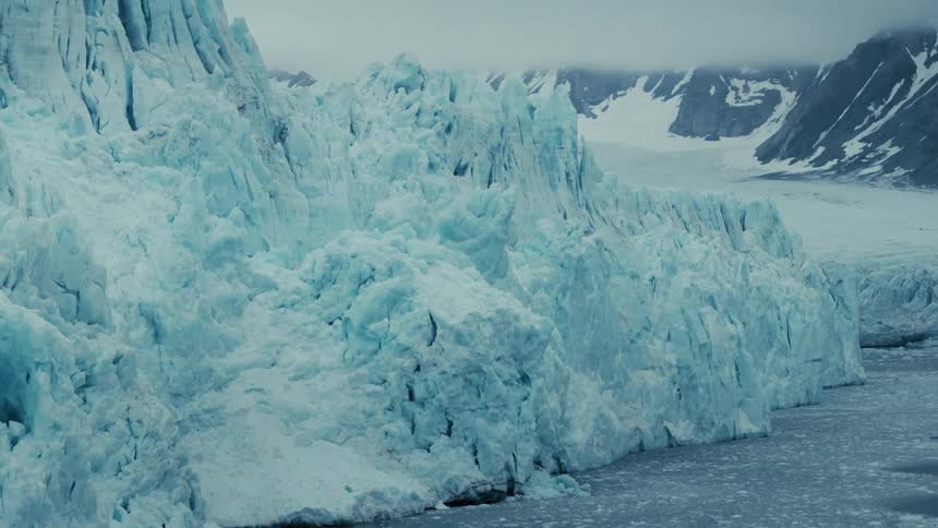A slow tilt reveals a majestic blue glacier cliff meeting the icy arctic sea, with snowy mountains in the distance