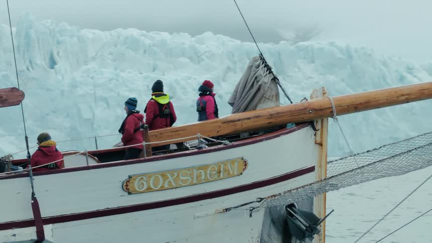 A group of tourists on a traditional wooden sailboat watch a massive glacier in the frigid waters of the Arctic