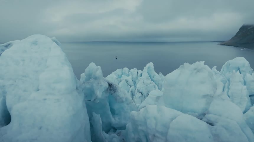A slow aerial shot pans over the immense, cracked surface of a glacier meeting the calm arctic ocean under an overcast sky