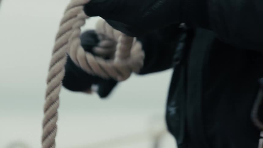 A sailor wearing black gloves methodically coils a thick rope on the deck of a boat