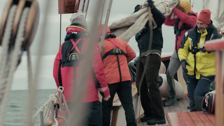 A crew of sailors in warm gear work together to stow a sail on the deck of a wooden ship on an overcast day