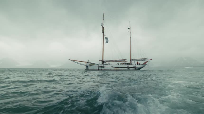 A classic two-masted sailboat is anchored in a remote, misty bay as the camera sails away, leaving a wake
