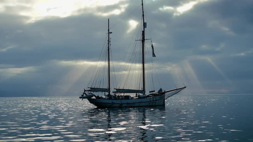 A classic sailboat drifts on a calm ocean as dramatic sunbeams break through the heavy clouds