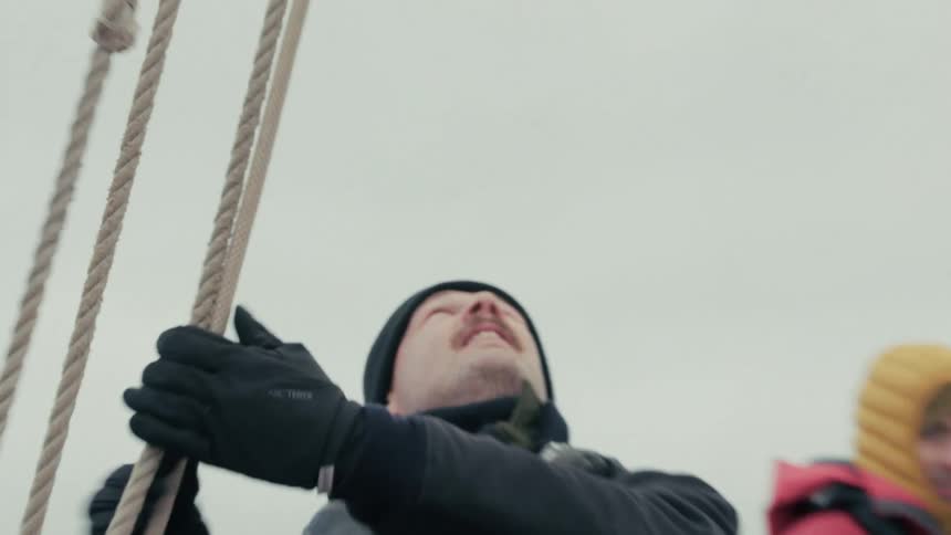 A sailor with a mustache wearing a black beanie and life vest pulls on the rigging of a ship