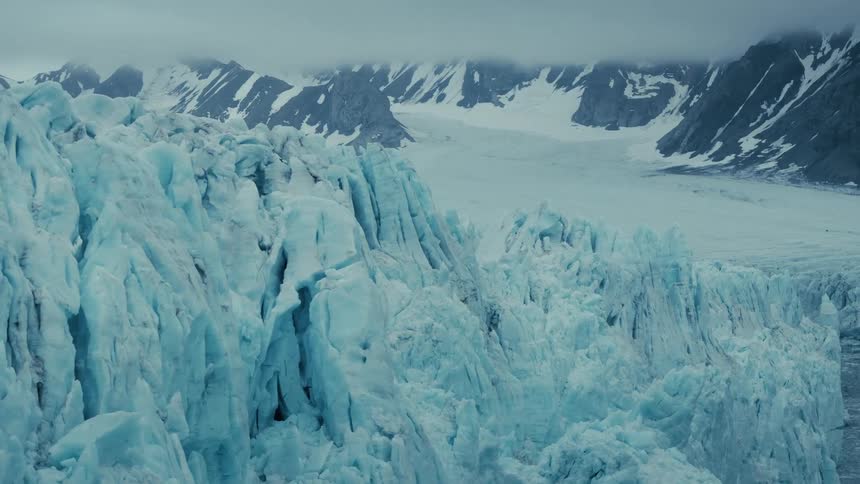 A majestic aerial view slowly tracks across the vast, crevassed face of a massive blue glacier nestled in a snowy mountain range