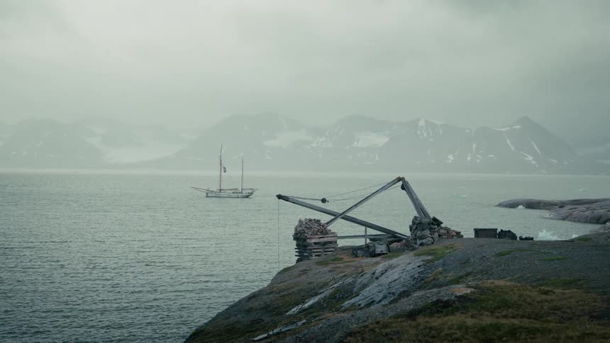 A rustic crane stands on a desolate cliffside overlooking a sailboat anchored in a foggy arctic bay with snowy mountains in the distance