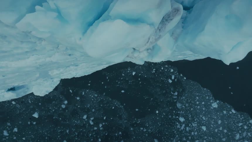 A vertical top-down aerial view slowly zooms out from a massive blue iceberg floating in the dark, cold arctic ocean