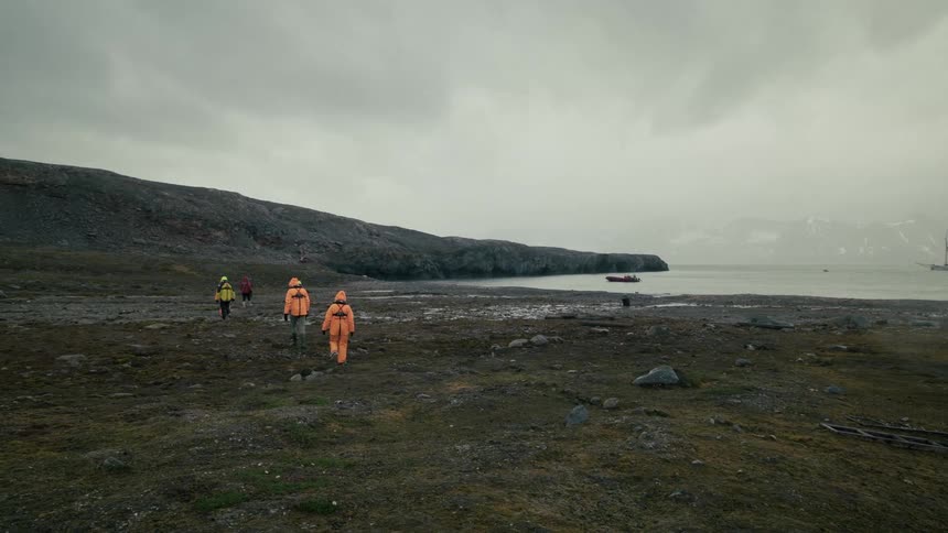 A group of explorers in orange suits walks across the desolate arctic tundra next to the ocean on an overcast day