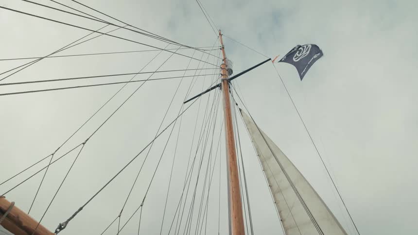 Slow tilt up a traditional wooden sailboat mast reveals a dark flag flapping in the wind against an overcast sky