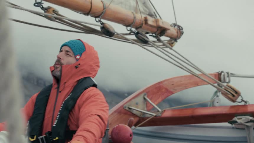 The captain of a wooden sailboat steers the vessel through a foggy and mountainous landscape