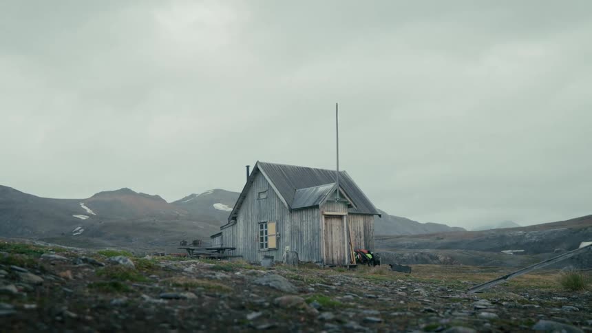 A weathered wooden cabin stands alone in a vast, desolate mountain landscape under an overcast sky