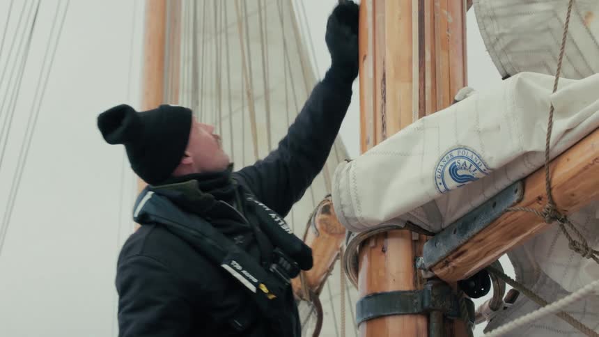 A sailor wearing a beanie and life vest hoists a sail on the wooden mast of a traditional tall ship