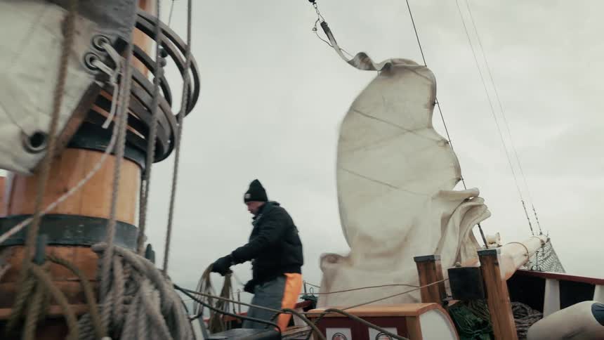 A sailor in foul weather gear coils a rope on the deck of a wooden ship in slow motion