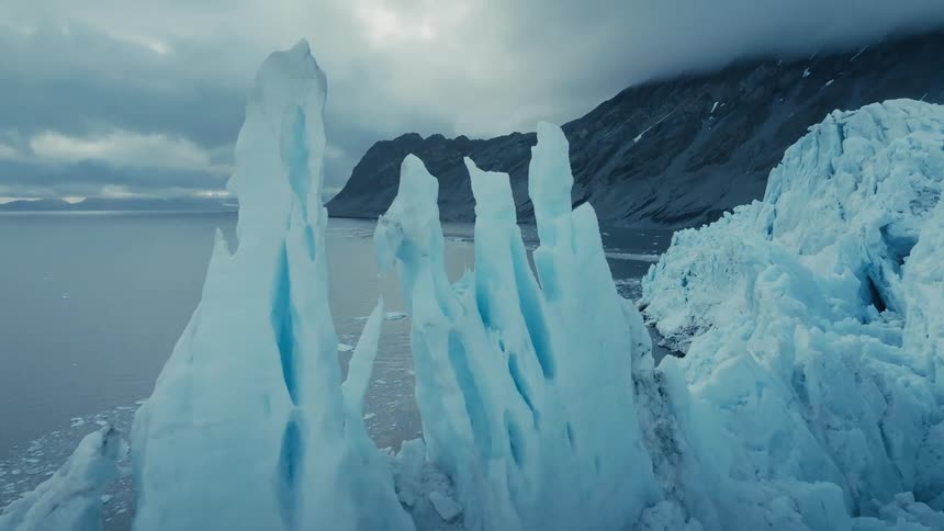 A dramatic slow aerial push through jagged blue glacial ice formations with the cold Arctic ocean and mountains in the background