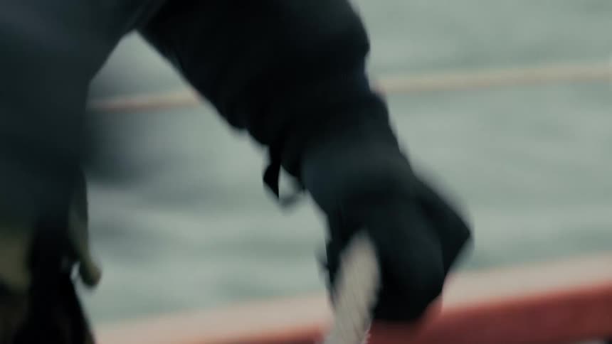 A person wearing black gloves pulls a thick rope hand over hand on the deck of a boat in slow motion