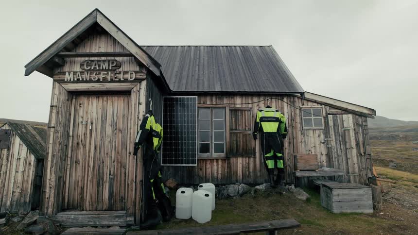 Rustic 'Camp Mansfield' wooden cabin with a solar panel and drying suits in a barren, remote landscape