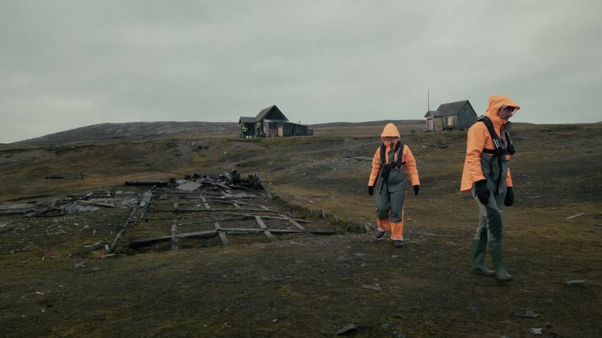 Slow motion shot of two explorers in bright orange outdoor gear walking through a bleak, abandoned arctic outpost