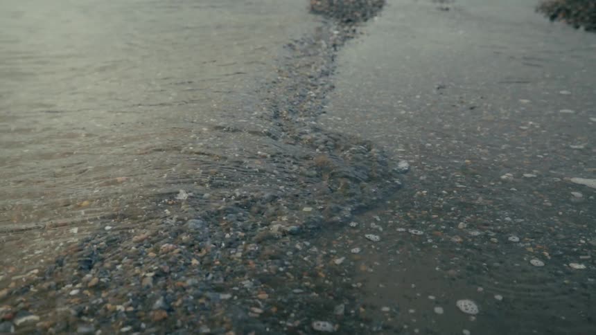 Slow motion closeup of calm, clear water gently flowing over a shoreline of small pebbles and sand