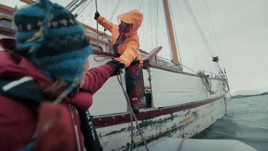 Slow motion over-the-shoulder shot of a sailor in a dinghy throwing a rope to a crewmate on a large wooden sailboat in arctic waters