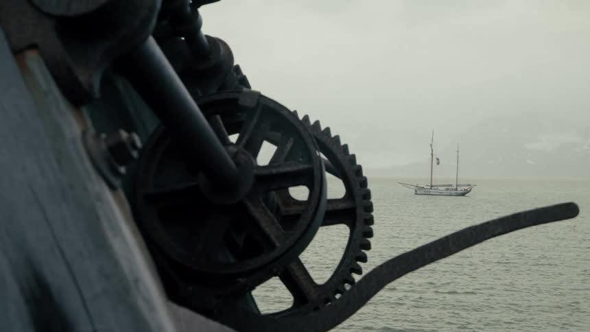 An old, rusty winch gear on a pier frames a vintage sailboat anchored in a misty bay