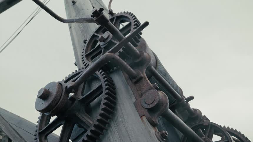 A slow zoom out of a complex, rusty antique industrial winch mechanism with intricate gears against an overcast sky