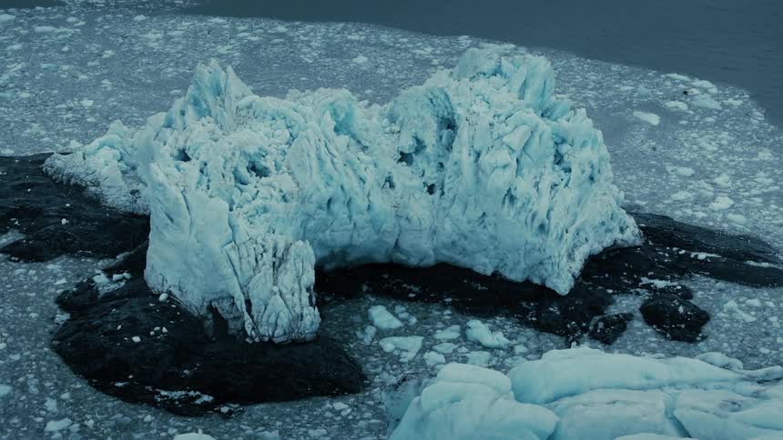 An enormous, jagged blue iceberg is grounded on dark rocks, surrounded by a sea of floating ice