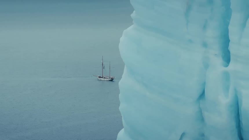 A classic two-masted sailboat glides through the tranquil arctic ocean, disappearing behind a towering blue iceberg