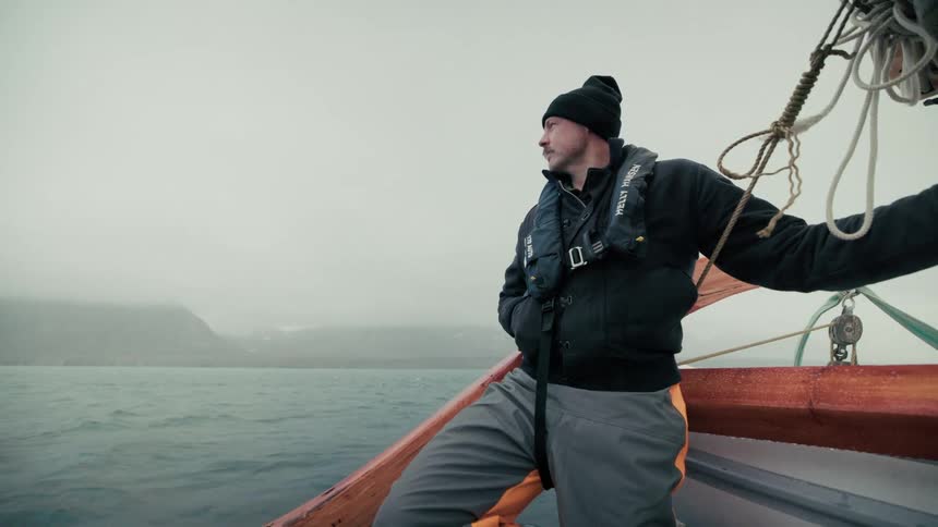 A man in a beanie and life vest stands on a boat, looking out over the cold, foggy sea towards a distant coastline