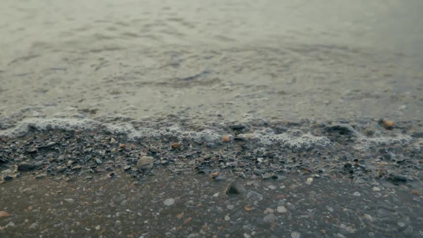 A serene, slow-motion close-up of a gentle wave lapping against a shore of small pebbles and shells