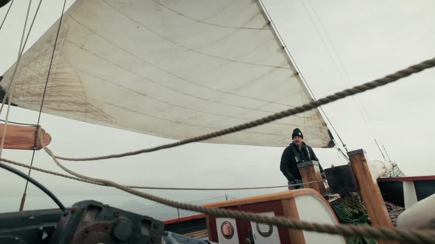 A sailor in a beanie pulls on a rope to adjust the large sail of a classic wooden boat on an overcast day