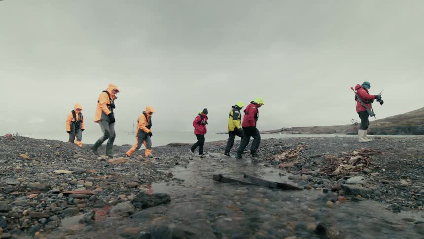 An expedition team in brightly colored winter gear walks in slow motion across a rocky arctic beach and a small stream
