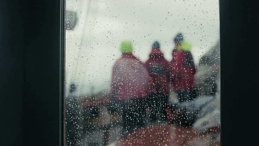 Sailors in red raincoats stand on the deck of a ship during a storm, seen through a rainy window