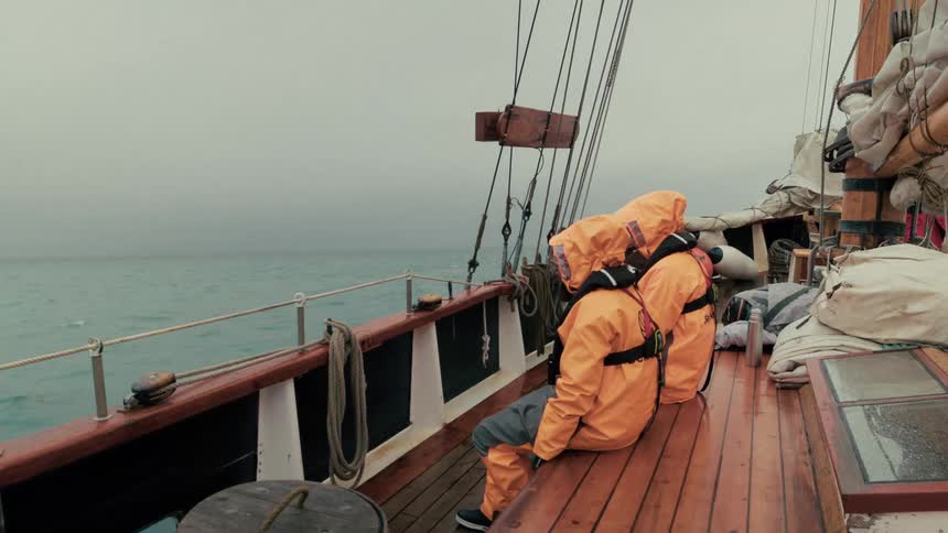 Two sailors in bright orange rain gear sit on the wooden deck of a moving sailboat on a cold, overcast day