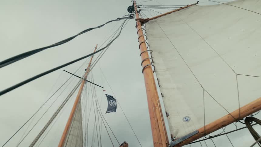 A low-angle view of the wooden mast and sails of a classic schooner as a flag flaps in the wind against a grey, overcast sky