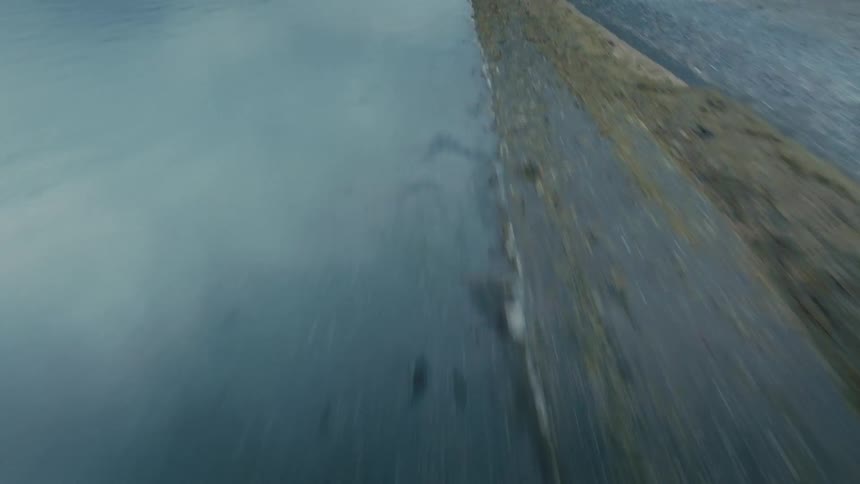 A high-speed FPV drone flies low over a barren coastline, tilting up to reveal a vast, dramatic landscape of mountains and water under a cloudy arctic sky