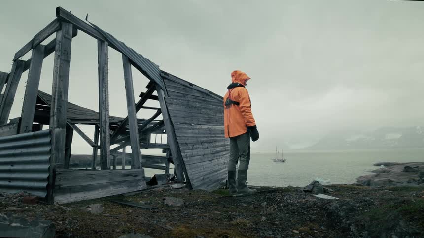 In slow motion, a solitary explorer in an orange jacket walks from a ruined coastal hut on a bleak, overcast day