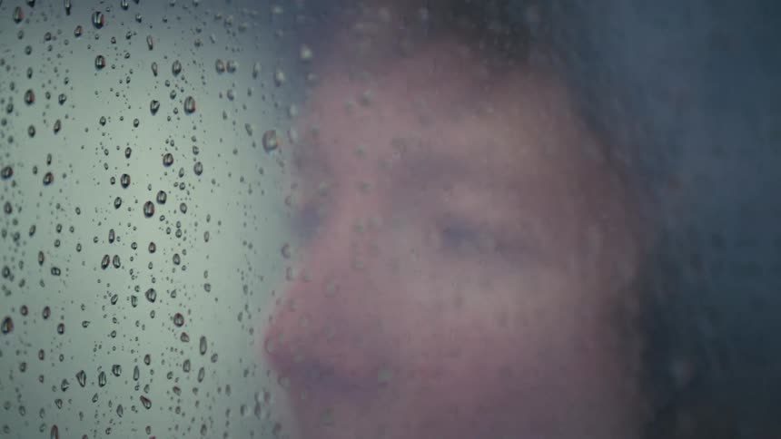 A sad woman closes her eyes as she looks out a window, with the focus pulling to the raindrops on the glass