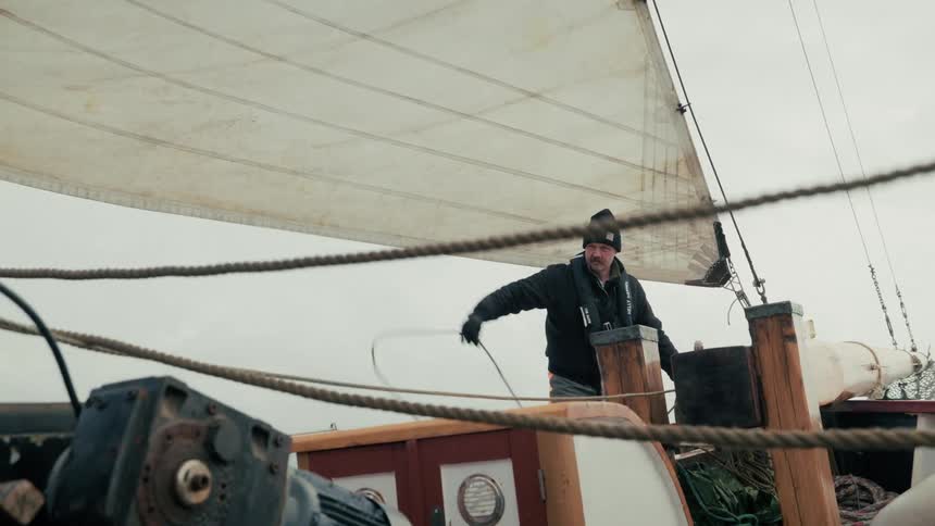 A sailor with a mustache trims the sail by pulling a rope on a traditional wooden boat under a grey, overcast sky