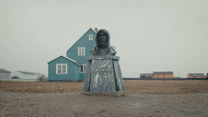 A stone monument of an explorer stands before a bright blue house in a remote Arctic village under an overcast sky