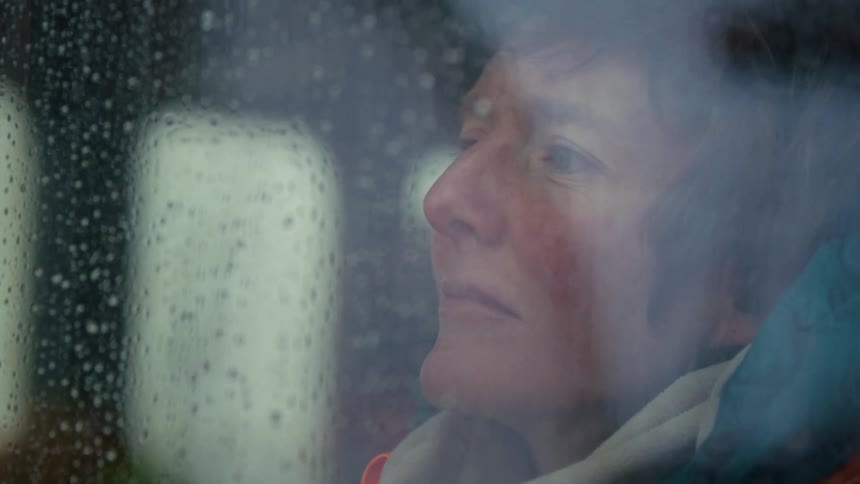 A pensive woman with a melancholic expression looks out a window as rain falls on the glass