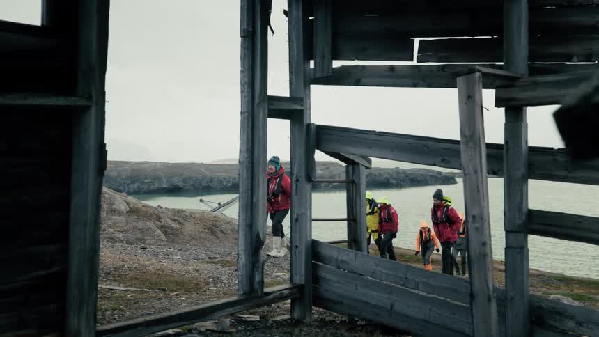 A group of arctic explorers walk along a remote coastline, framed by the ruins of an old wooden building