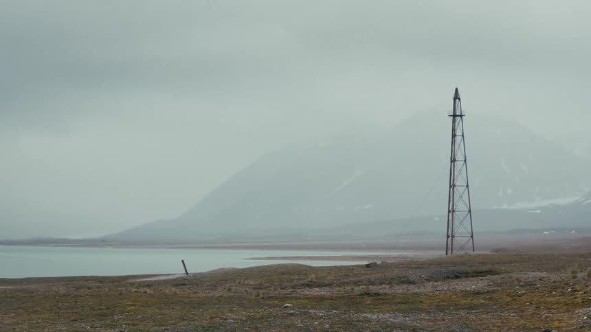 A desolate and foggy landscape shows a lone pylon standing by a calm lake with snow-dusted mountains in the distance