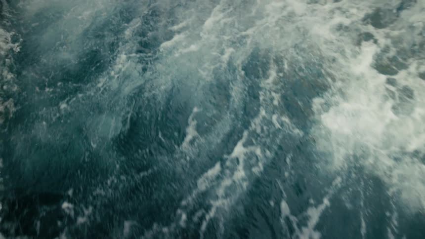 A high-angle view captures the side of a boat as it cruises through the water, tilting down to focus on the churning, foamy wake
