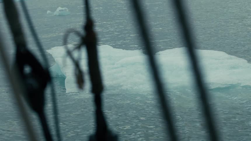 An iceberg drifts through the cold ocean water as seen from the deck of a sailing ship