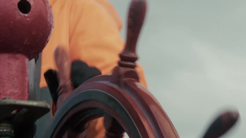 A sailor in an orange jacket rapidly spins the wooden steering wheel of a ship at sea